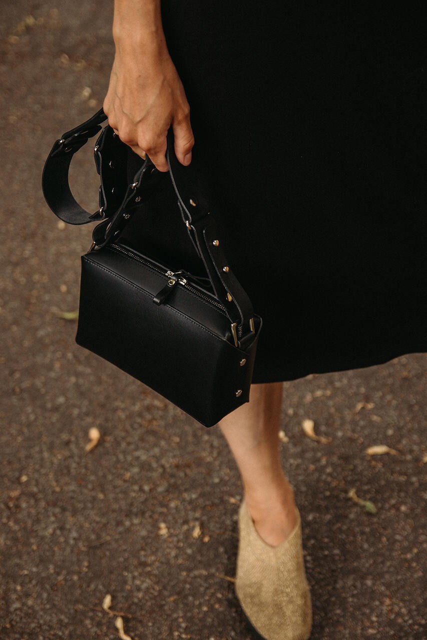 Sac bandoulière en cuir noir Stella Soomlais, tenu à la main, dessus zippé et sangle rivetée