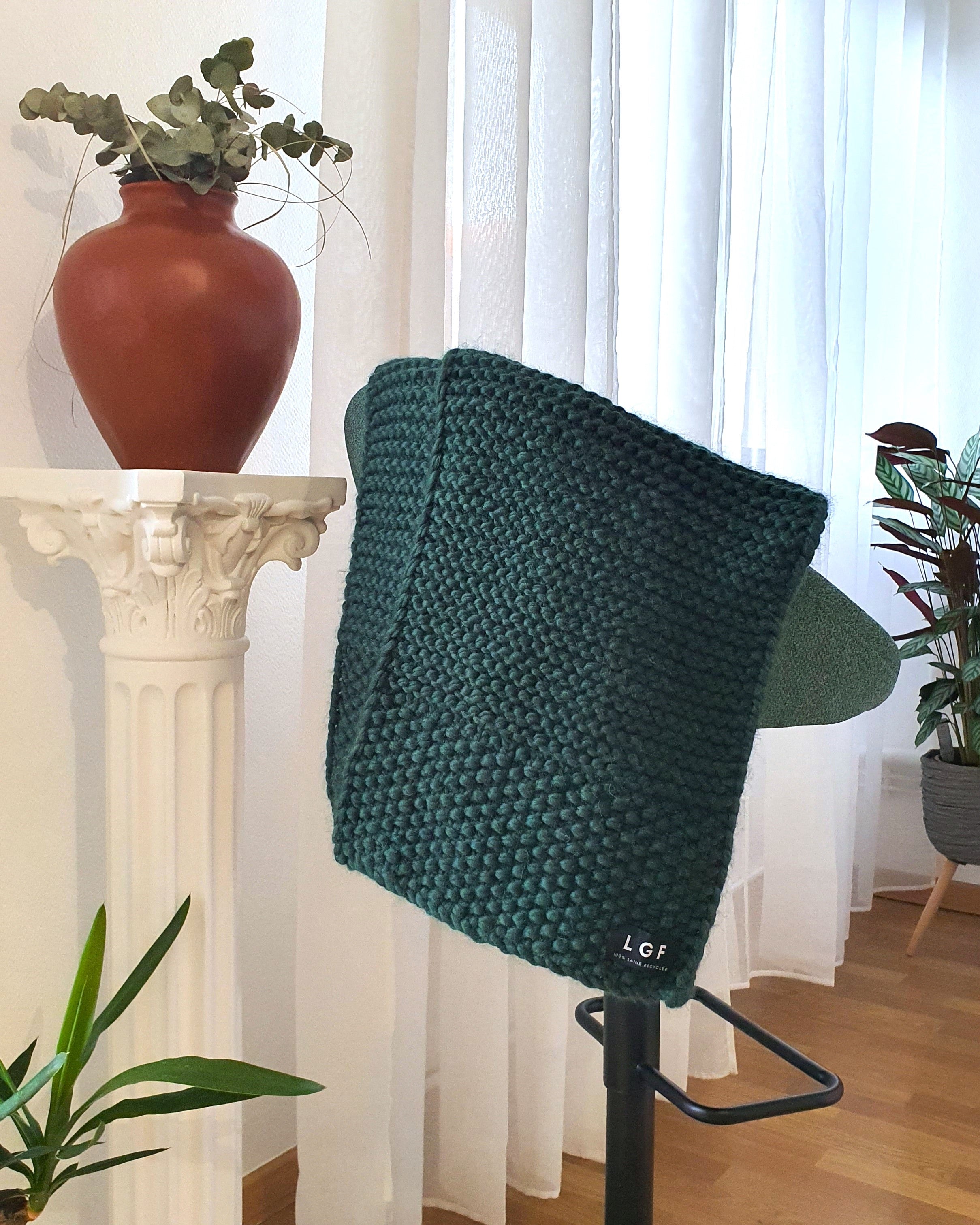 Plaid en grosse maille de laine point mousse vert sapin, drapé sur un mannequin dans un intérieur décoré