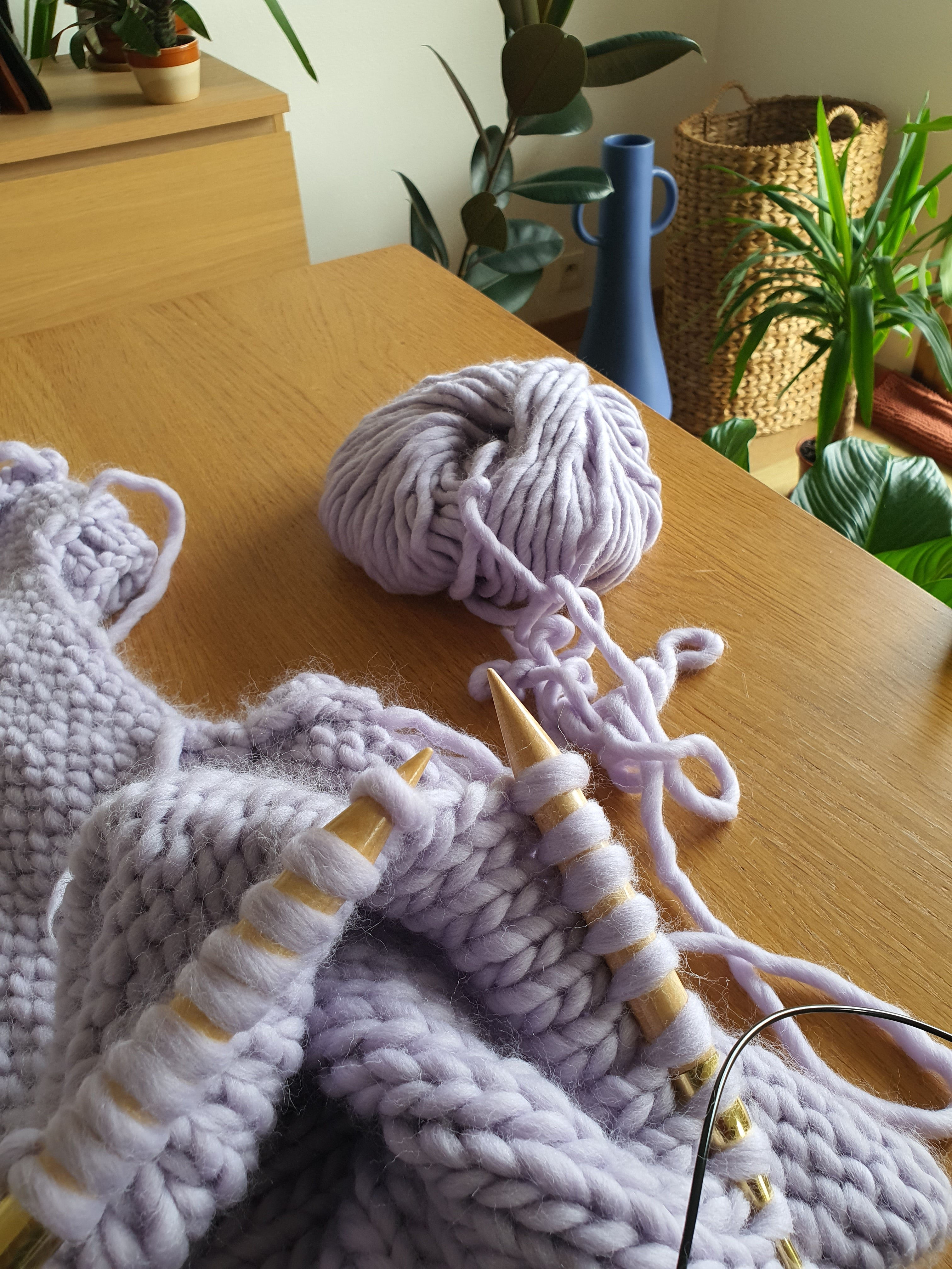 Pelote de laine épaisse lilas avec aiguilles à tricoter sur une table en bois, plantes en pot et vase bleu à côté