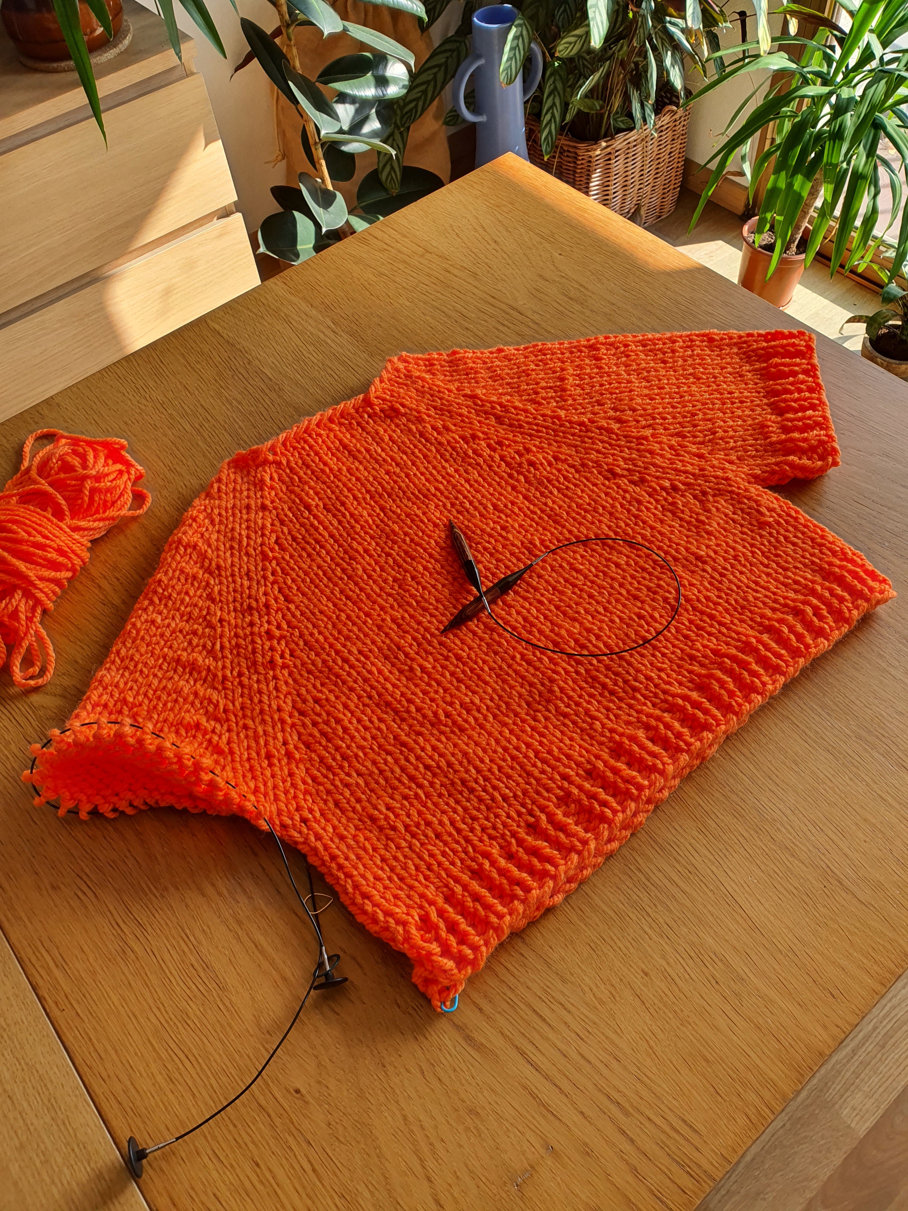 Pull en laine orange corail porté en intérieur avec écheveaux de laine assortis posés autour