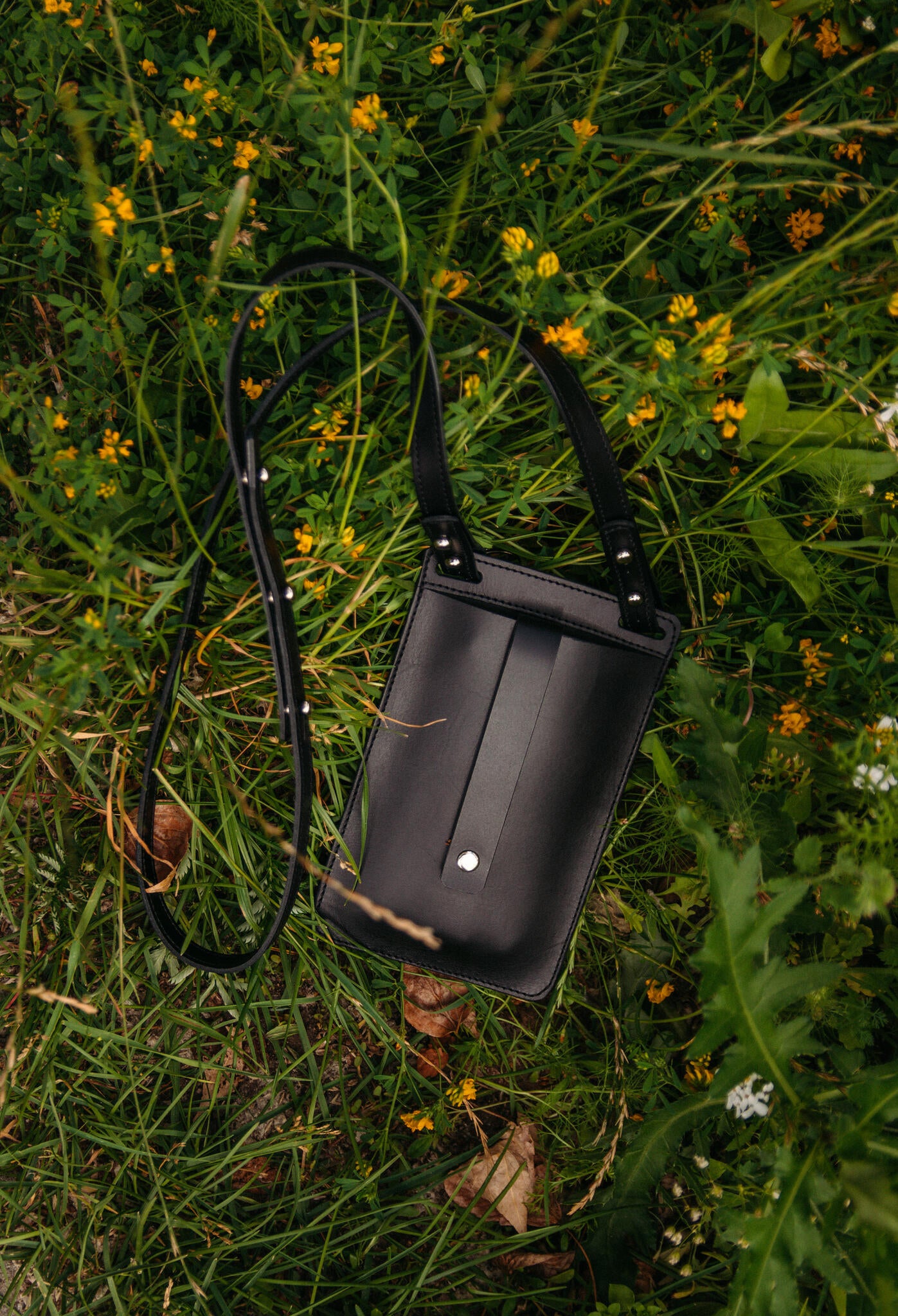 Pochette téléphone en cuir noir Stella Soomlais à plat parmi les fleurs jaunes, bandoulière réglable déployée
