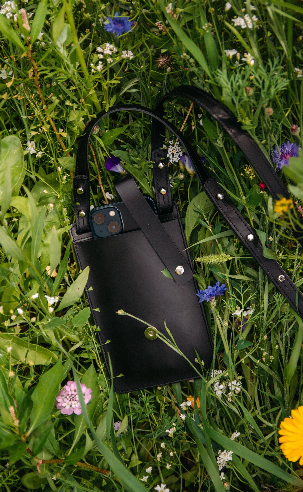 Pochette téléphone en cuir noir Stella Soomlais parmi bleuets et fleurs sauvages, téléphone dépassant de la poche