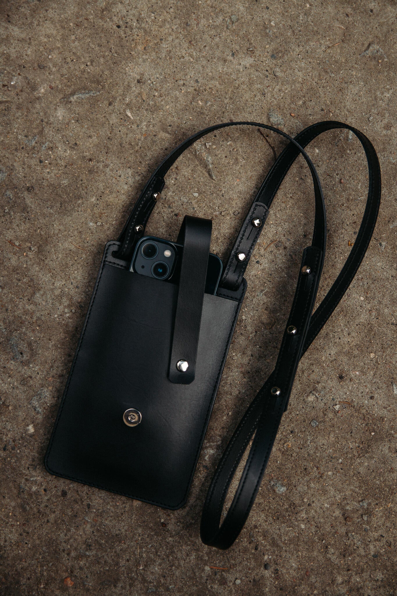 Pochette téléphone en cuir noir Stella Soomlais sur béton, smartphone rangé dépassant de l'ouverture