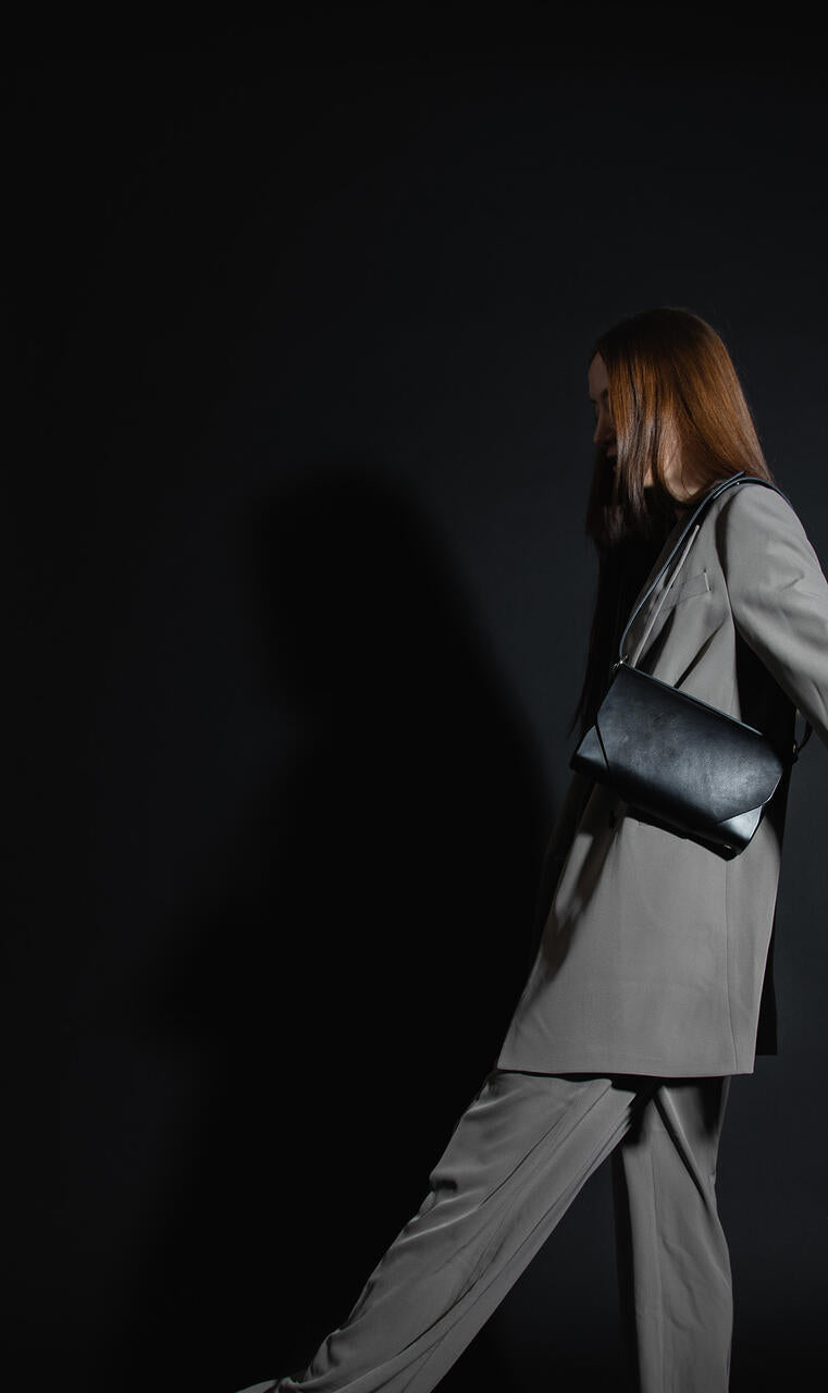 Sac bandoulière en cuir noir Stella Soomlais porté à l'épaule par un modèle en tailleur gris