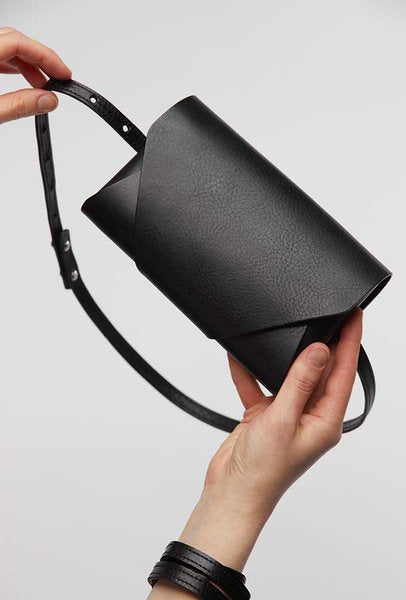 Pochette-portefeuille en cuir noir Stella Soomlais tenue en mains, rabat plié et bandoulière ajustable rivetée