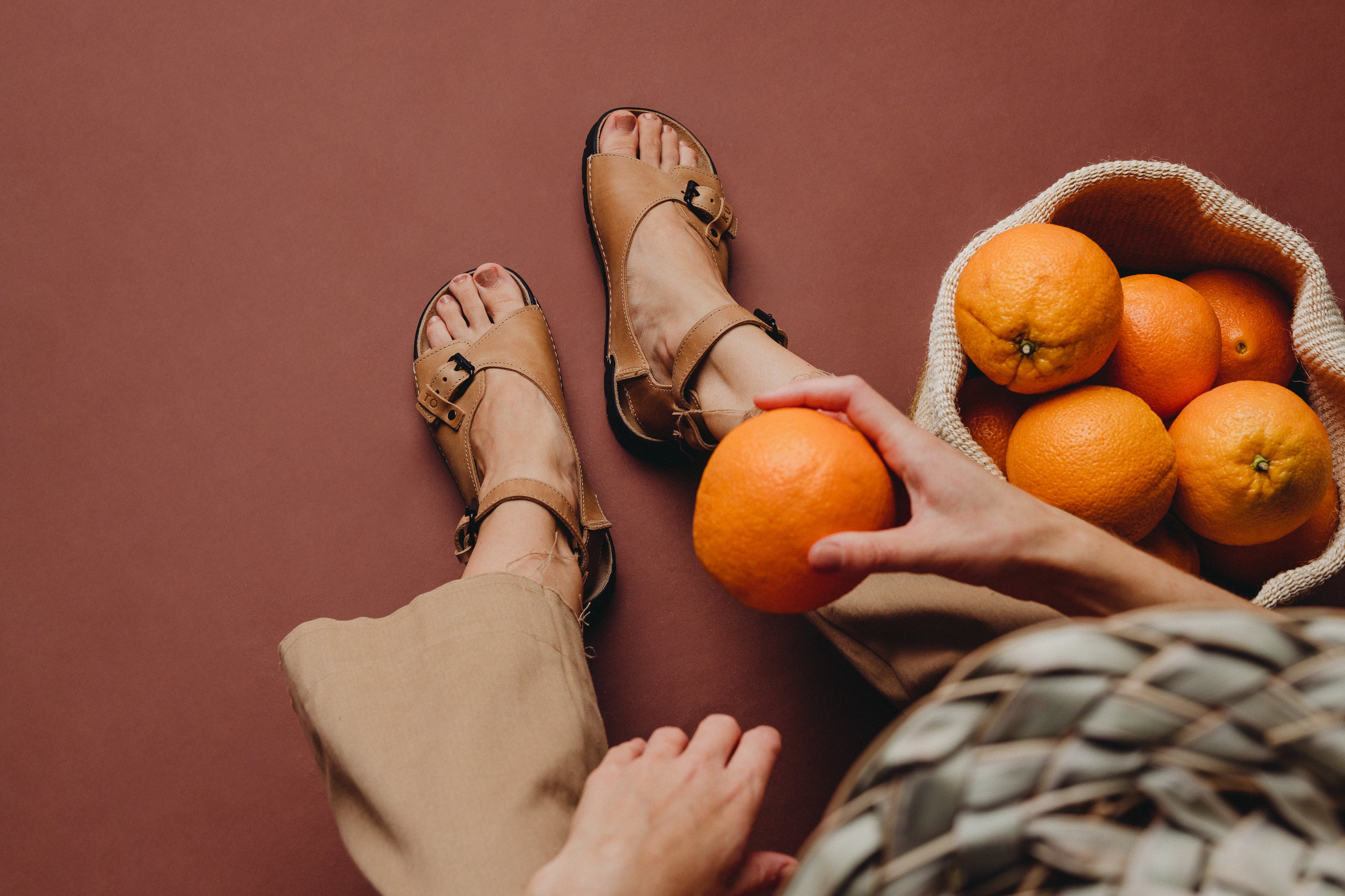 Sandales en cuir beige tenant une orange, panier d'oranges en arrière-plan