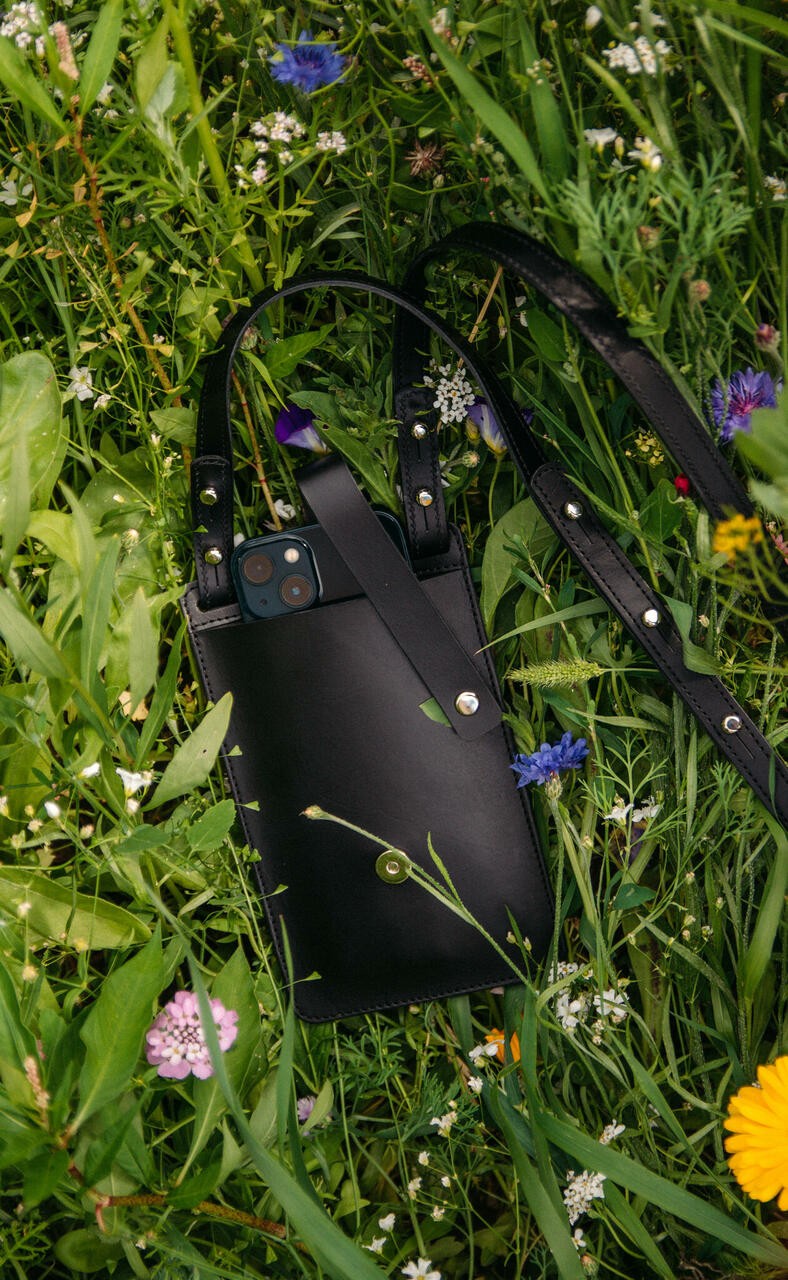 Pochette téléphone en cuir noir Stella Soomlais dans une prairie fleurie, smartphone glissé à l'intérieur