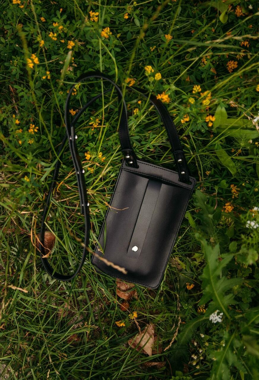 Pochette téléphone en cuir noir Stella Soomlais posée dans l'herbe fleurie, poche avant et bouton pression