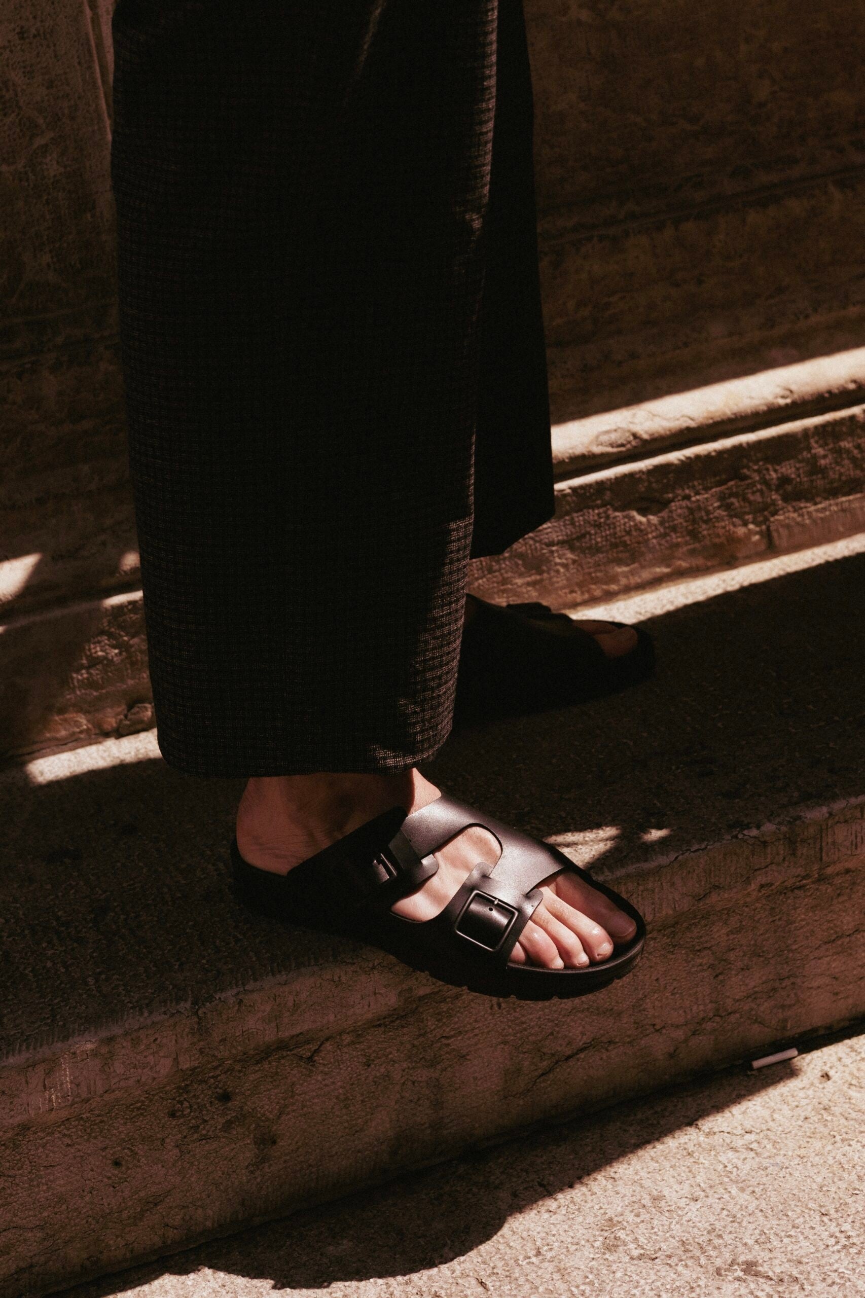 Pieds d’un homme en pantalon foncé portant des sandales TOKU Zurich en cuir noir à deux brides à boucles, descendant des marches en pierre au soleil