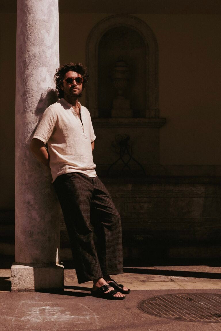 Homme en chemise de lin claire portant des sandales TOKU Zurich en cuir noir, adossé à une colonne de pierre près d’une fontaine ancienne, ambiance estivale