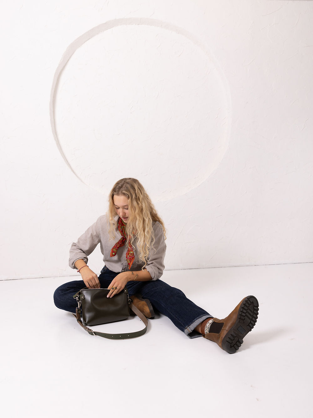 Modèle blonde aux cheveux longs assise au sol, chemise lin grise et foulard rouge paisley, jean brut et bottines marron, sac TOMMY en cuir vert thym posé entre les jambes, lookbook studio cercle peint, signature éditoriale TOKS VISOKS