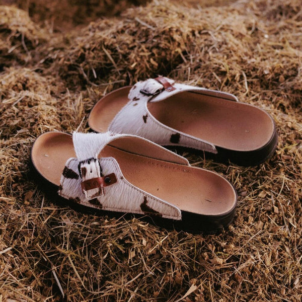 Paire de sandales TOKU Rome en cuir à poil imprimé vache posée sur du foin
