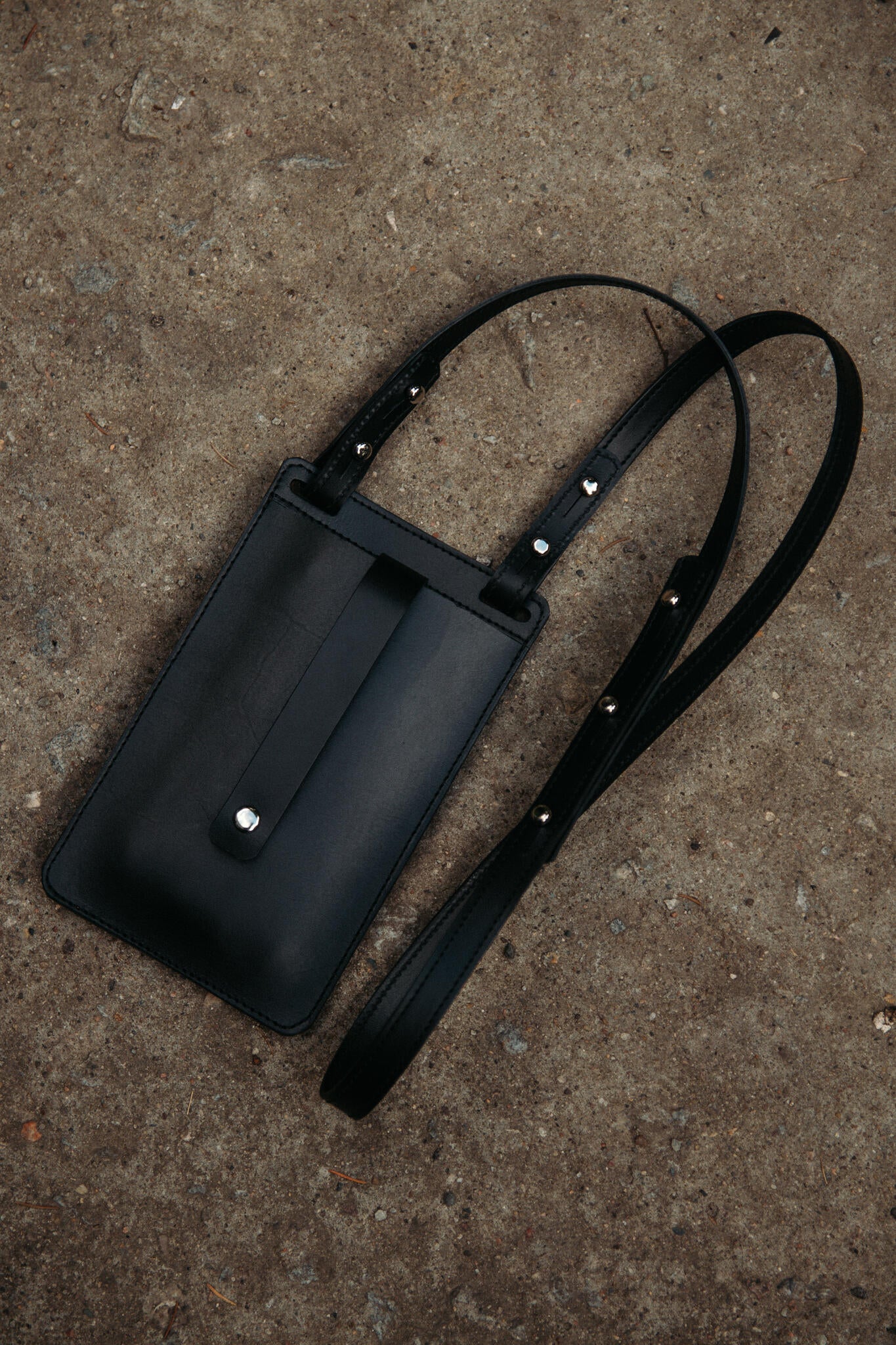 Pochette téléphone en cuir noir Stella Soomlais posée sur béton, vue de face avec patte et bouton pression nickel