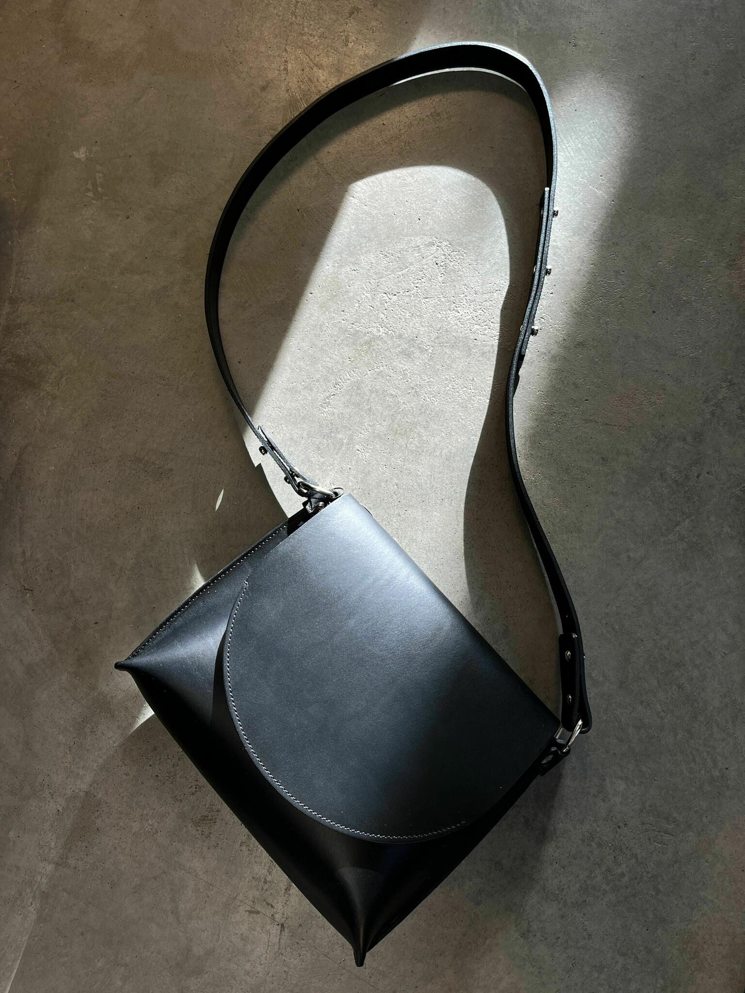Sac à main en cuir noir Stella Soomlais posé à plat, rabat arrondi et large sangle réglable