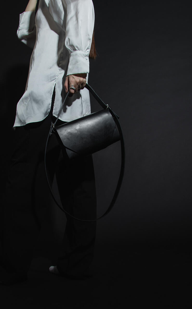 Sac bandoulière en cuir noir Stella Soomlais tenu à bout de bras par sa longue sangle