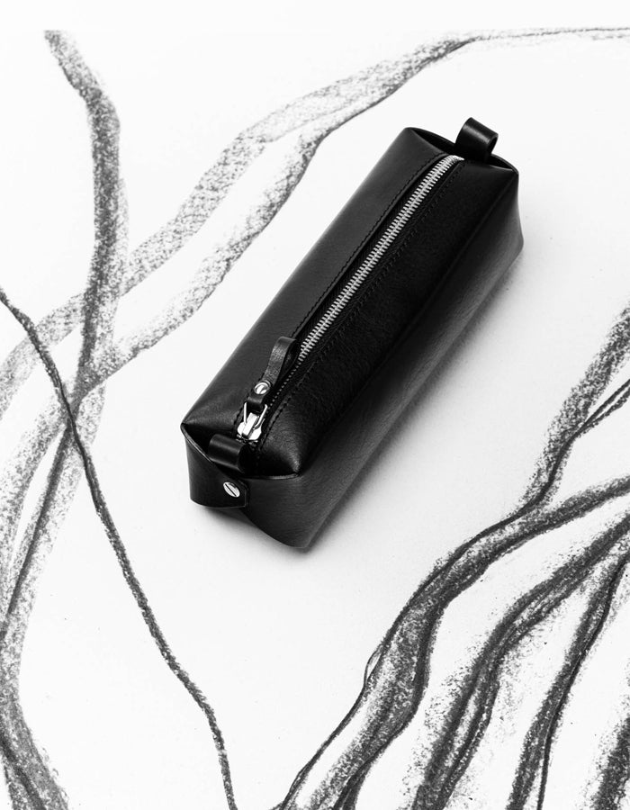 Trousse en cuir noir Single-Decker Stella Soomlais, posée à plat sur un fond dessiné au fusain