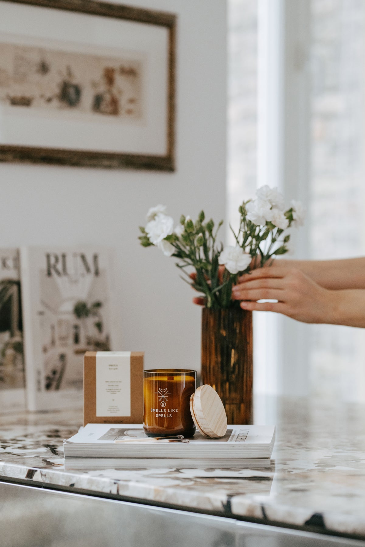 Bougie parfumée en verre ambré Smells Like Spells sur table en marbre, boîte et vase à côté