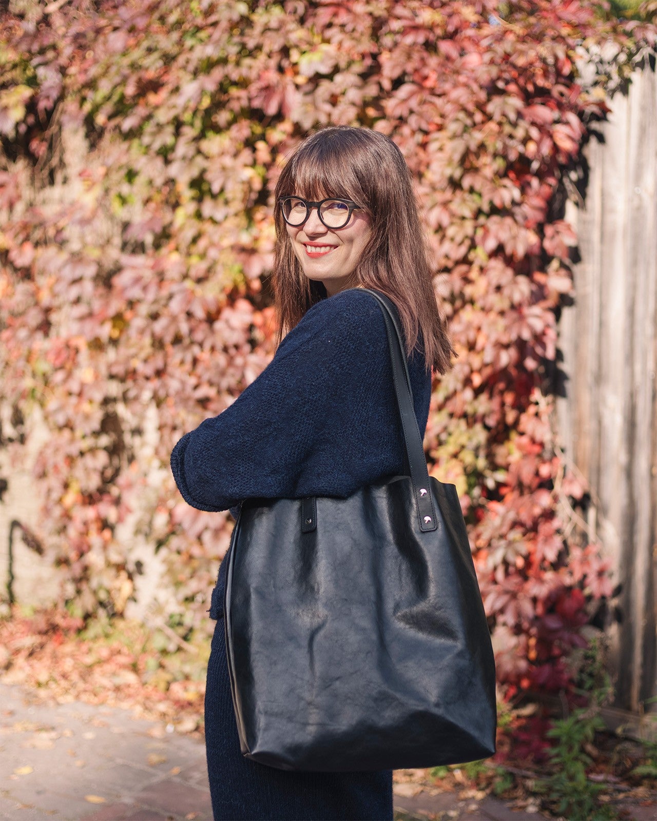 Sac cabas en cuir noir Stella Soomlais, porté à l'épaule devant un feuillage d'automne
