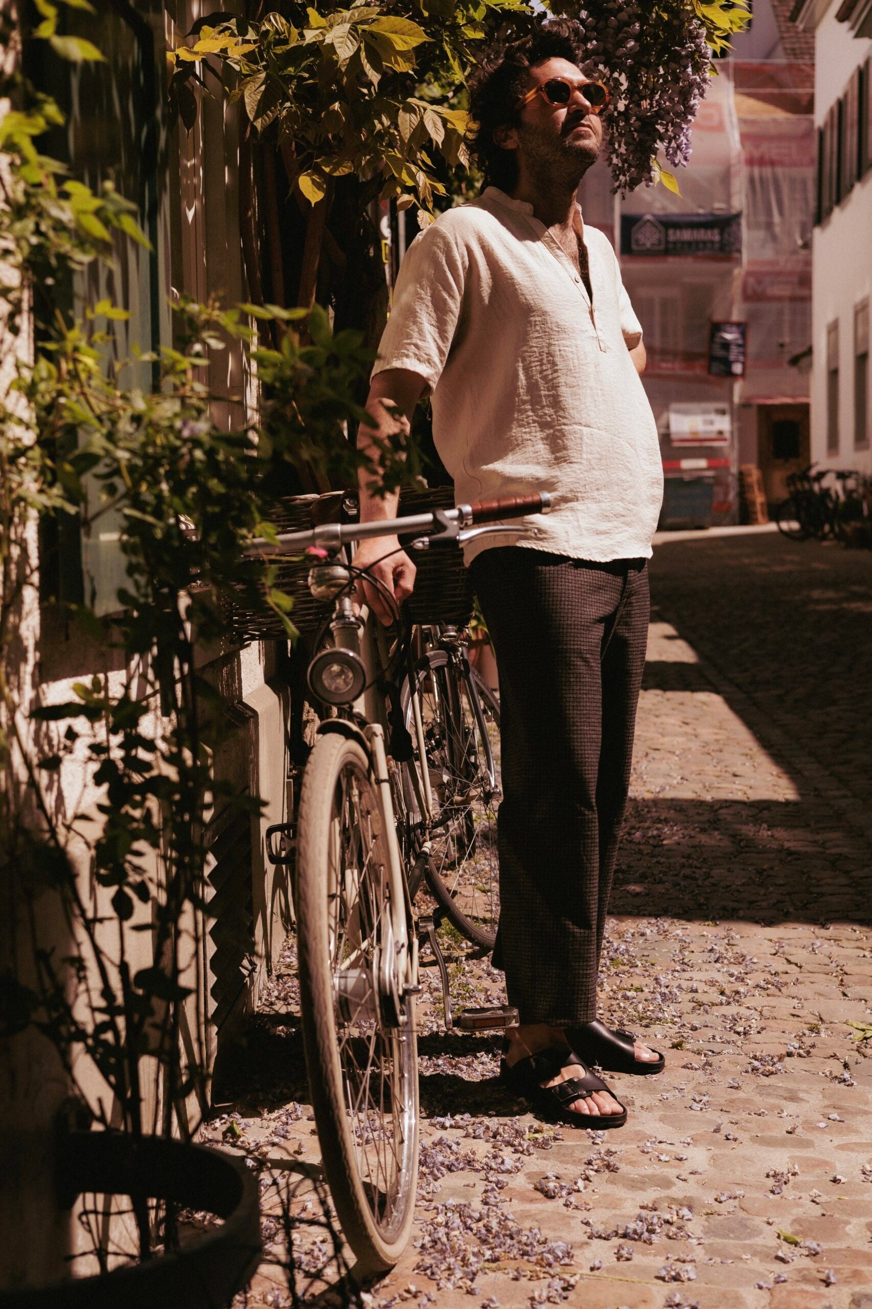 Homme en chemise de lin claire et pantalon foncé portant des sandales TOKU Zurich en cuir noir à deux brides, debout avec un vélo vintage dans une ruelle pavée ensoleillée